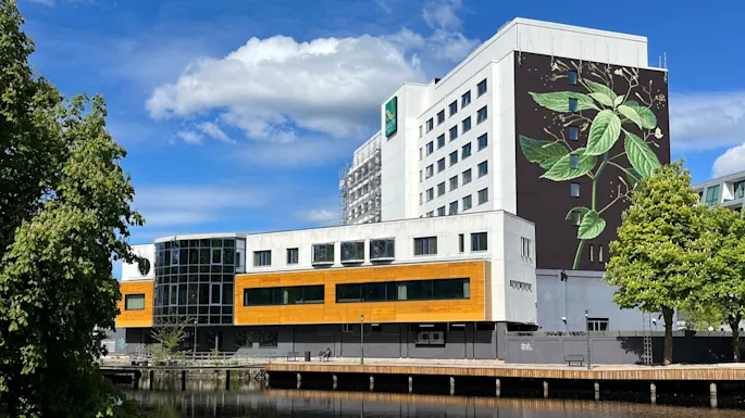 En fler-etasjes bygning viser et stort botanisk veggmaleri på fasaden. Den står ved siden av et rolig vassdrag, omgitt av frodige grønne trær, under en klar blå himmel. Tekst: "SCANDIC".