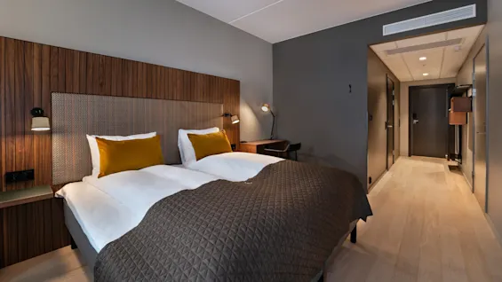 En dobbeltseng med hvitt sengetøy og gule puter står i et moderne hotellrom. Tregulv og vegger omgir et skrivebord med lamper og leder til en korridor.