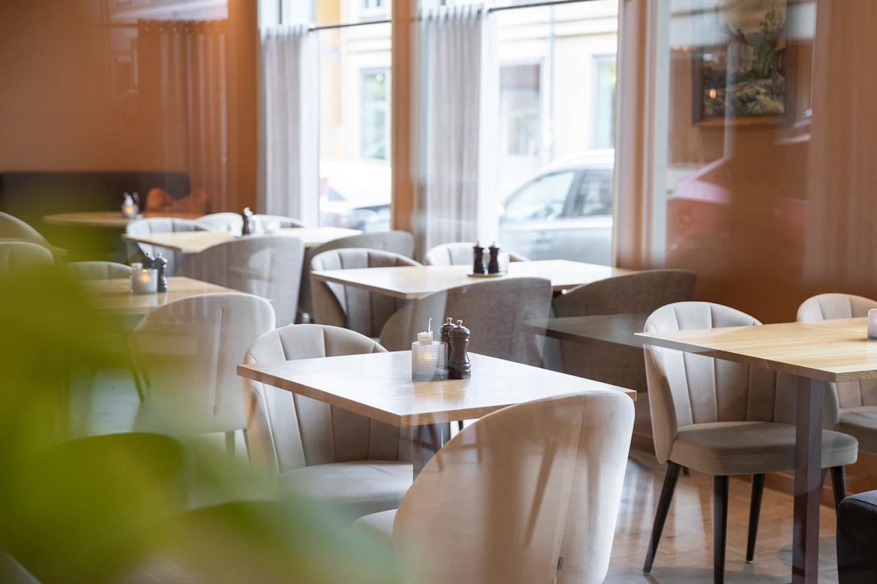 Bord og stoler er pent plassert i en koselig kafé med dempet belysning. Store vinduer slipper inn naturlig lys, med utsikt over parkerte biler utenfor.