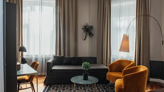 Et komfortabelt og stilfuldt værelse med en mørk sofa, to livlige lænestole og et skrivebord ved solbeskinnede vinduer.