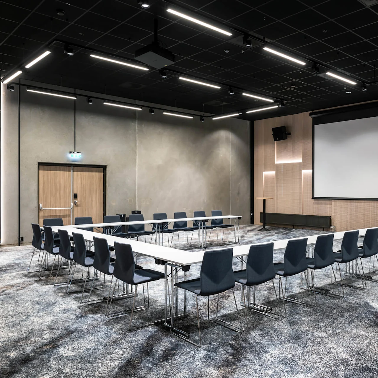 Konferencedel med U-formet bordopstilling, omgivet af marineblå stole. Projektorskræm på højre væg, abstrakt vægmaleri til venstre. Loftlys oplyser det moderne, tæppebelagte rum.