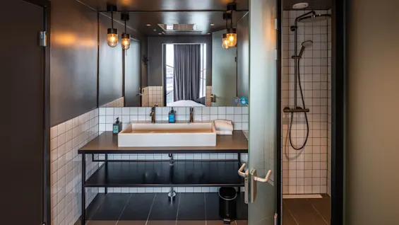 Et moderne bad med to vasker står under to hengende lys. Et speil reflekterer en delvis synlig seng. Til høyre står et flislagt dusjområde med glassdør åpen.