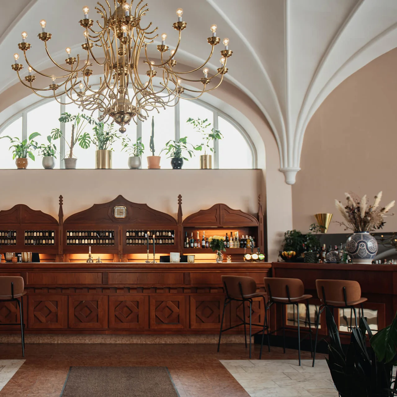 A wooden bar with high stools stands under an ornate chandelier. Shelves behind display bottles and plants adorn the upper ledge. The ceiling features elegant arches.