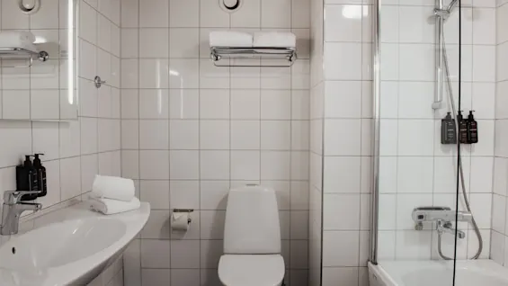 A clean, modern bathroom with white tiled walls, featuring a sink, toilet, and a shower with a glass screen. Towels are neatly folded.