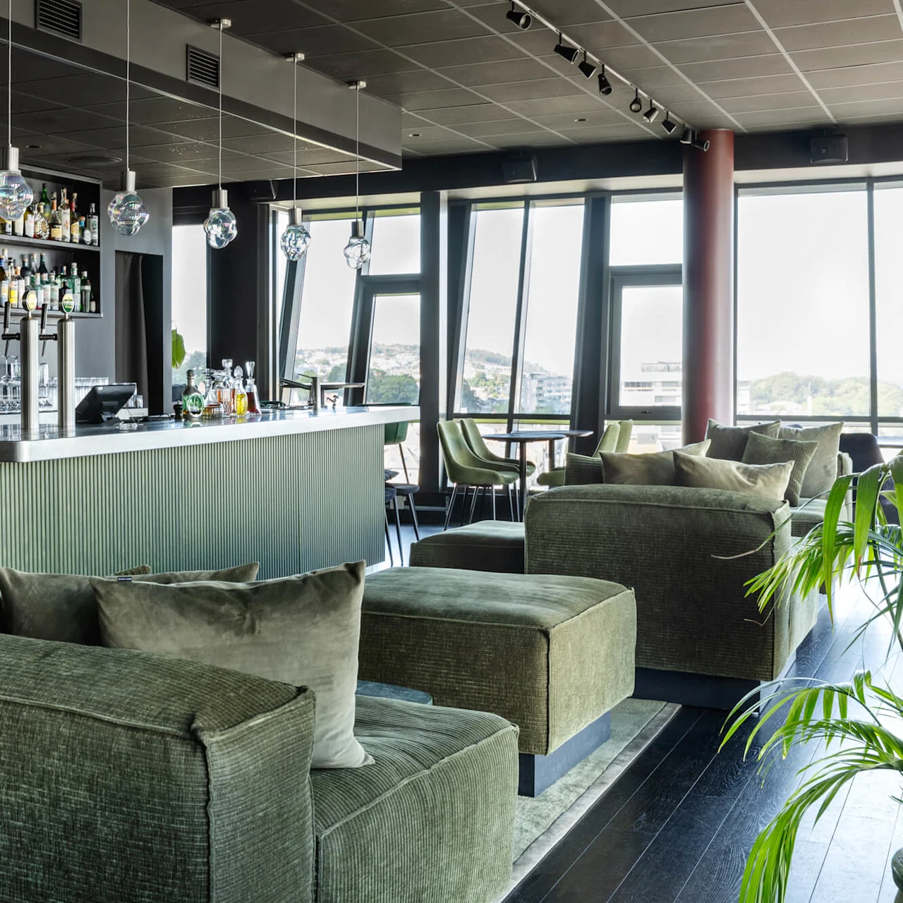 A modern bar features green upholstered seating, a sleek counter with hanging lights, and shelves stocked with bottles. Large windows provide a view of a distant cityscape.