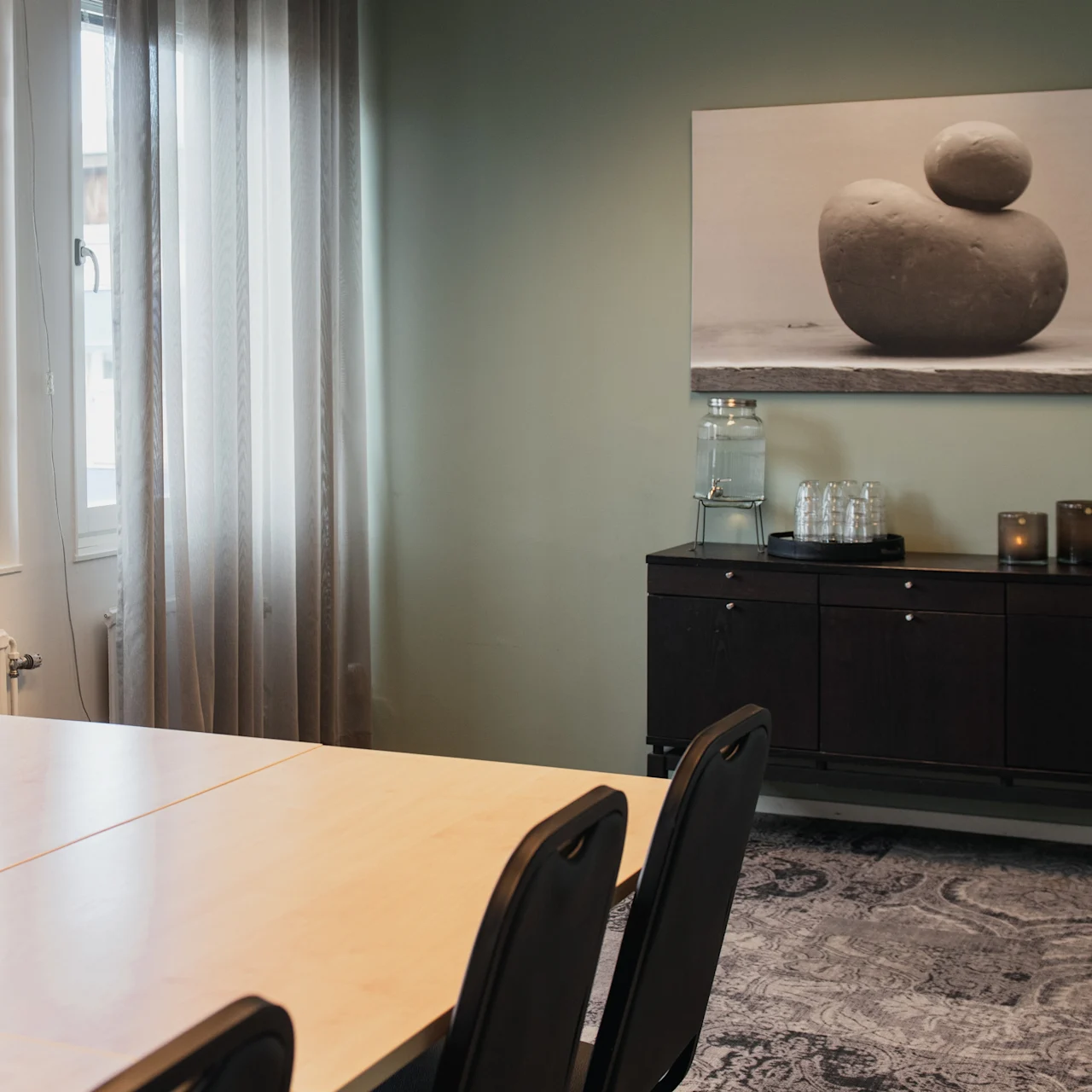 A wooden conference table with black chairs, situated in a room with green walls. A painting of stacked stones hangs above a black sideboard with a water dispenser.