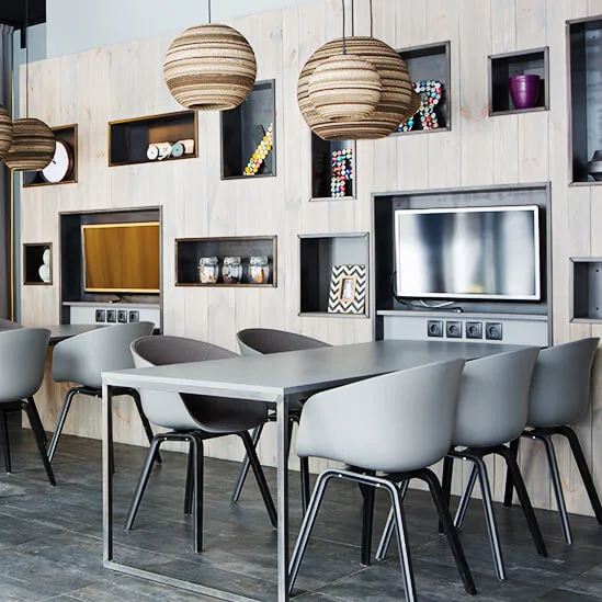 Tables with gray chairs sit in a modern room featuring pendant lights, decorated shelves, and a wall-mounted TV. Large windows with geometric patterns allow natural light to filter through curtains.