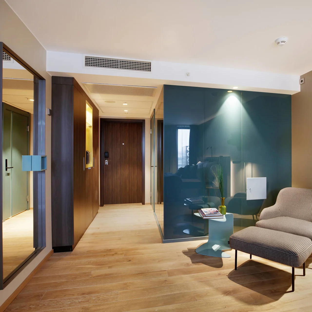 A lounge chair with footstool sits under a wall-mounted bear sculpture wearing glasses. A small side table holds magazines near a floor lamp on wooden flooring.