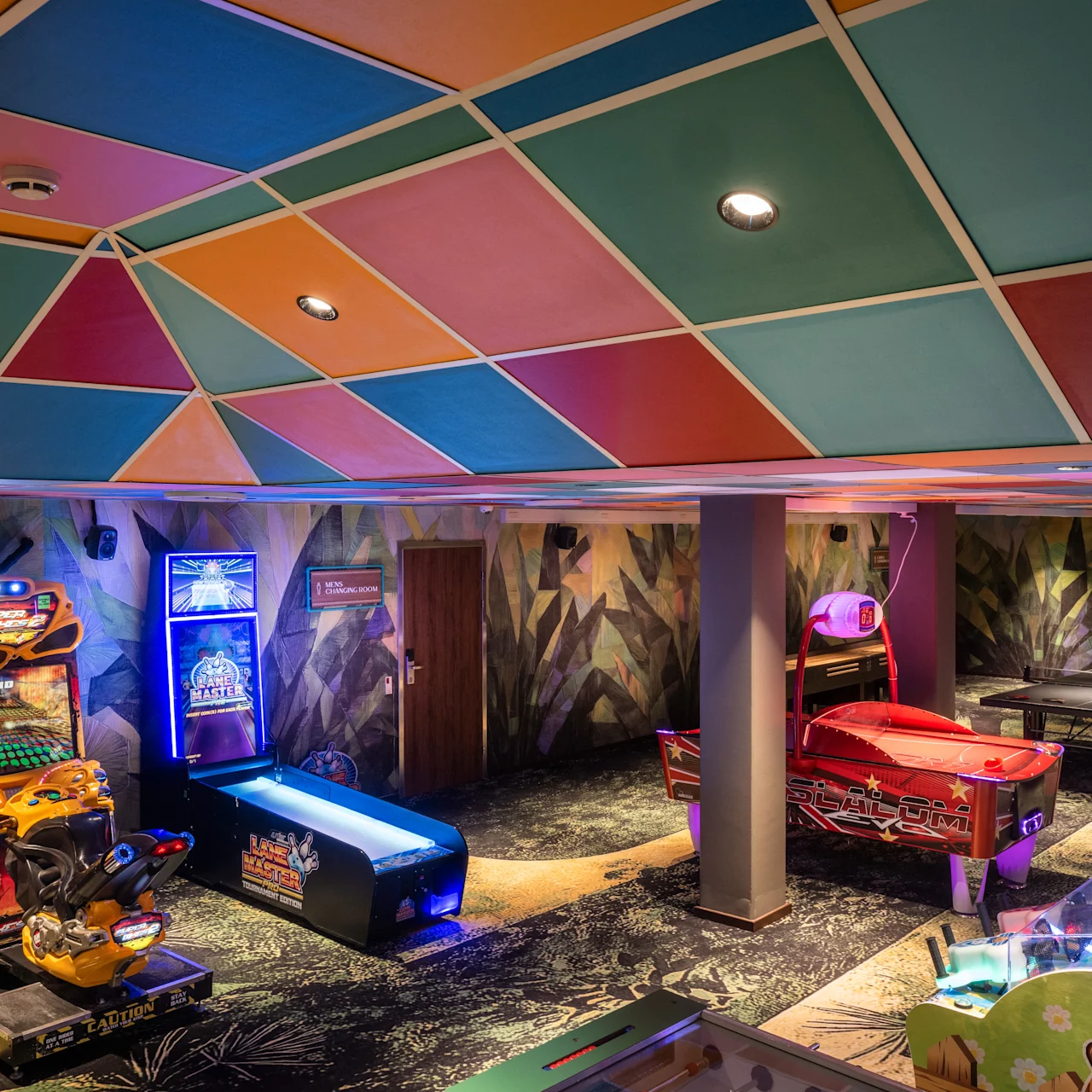 Arcade games stand in a colorful room, including racing games, air hockey, and pinball machines, under a vibrant, patterned ceiling. Neon lights illuminate the space, creating an exciting gaming environment.