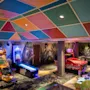 Arkadespil står i et farverigt rum, inklusive racerspil, airhockey og flipperspil, under et livligt, mønstret loft. Neonlys oplyser rummet og skaber en spændende spilatmosfære.