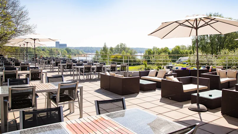 Outdoor seating area arranged with tables, chairs, and umbrellas, overlooking lush greenery and a distant building, under a clear blue sky. 