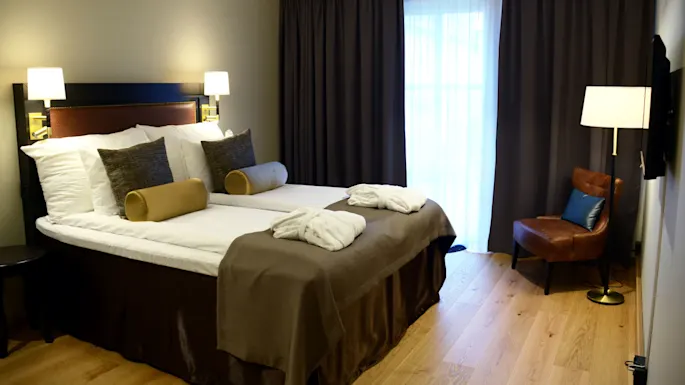 En pent oppredd seng med puter, i et godt opplyst hotellrom med gardiner, en stol og nattbordlamper.