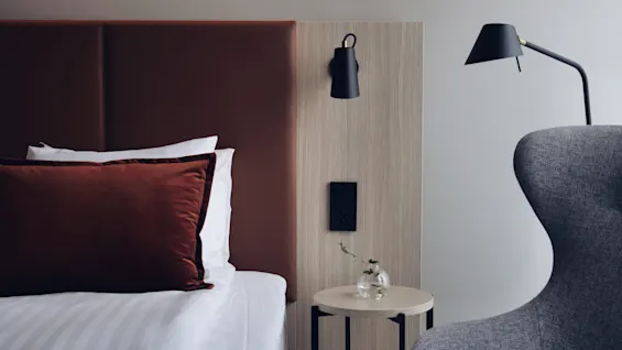 Rauhallinen hotellihuoneen sisustus siististi pedatulla sängyllä, pienellä sivupöydällä kasvin kera ja pehmeällä nojatuolilla rentoutumiseen.