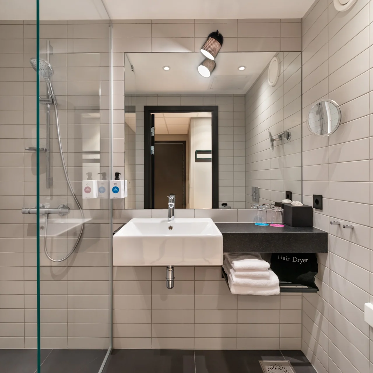 Et bad har en veggmontert vask under et speil med overlys. En glassdusj er ved siden av vasken. Teksten inkluderer "Hair Dryer" på en hylle med foldede håndklær.