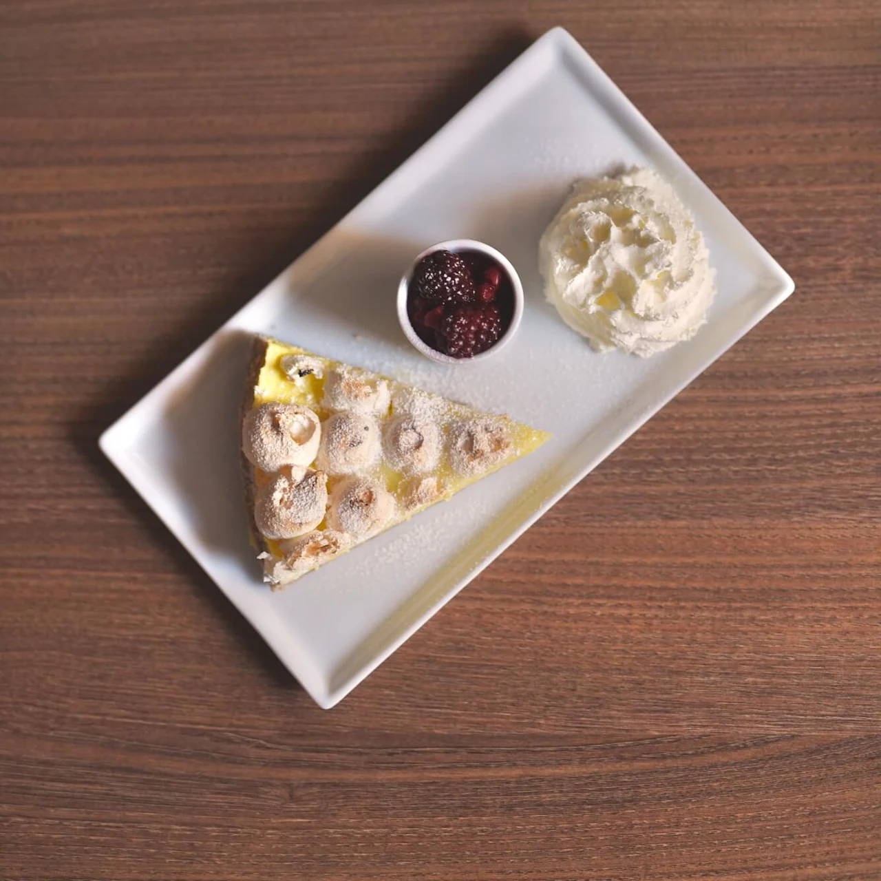 A slice of pie with powdered sugar, served with whipped cream and berry compote on a square plate, on a wooden table.