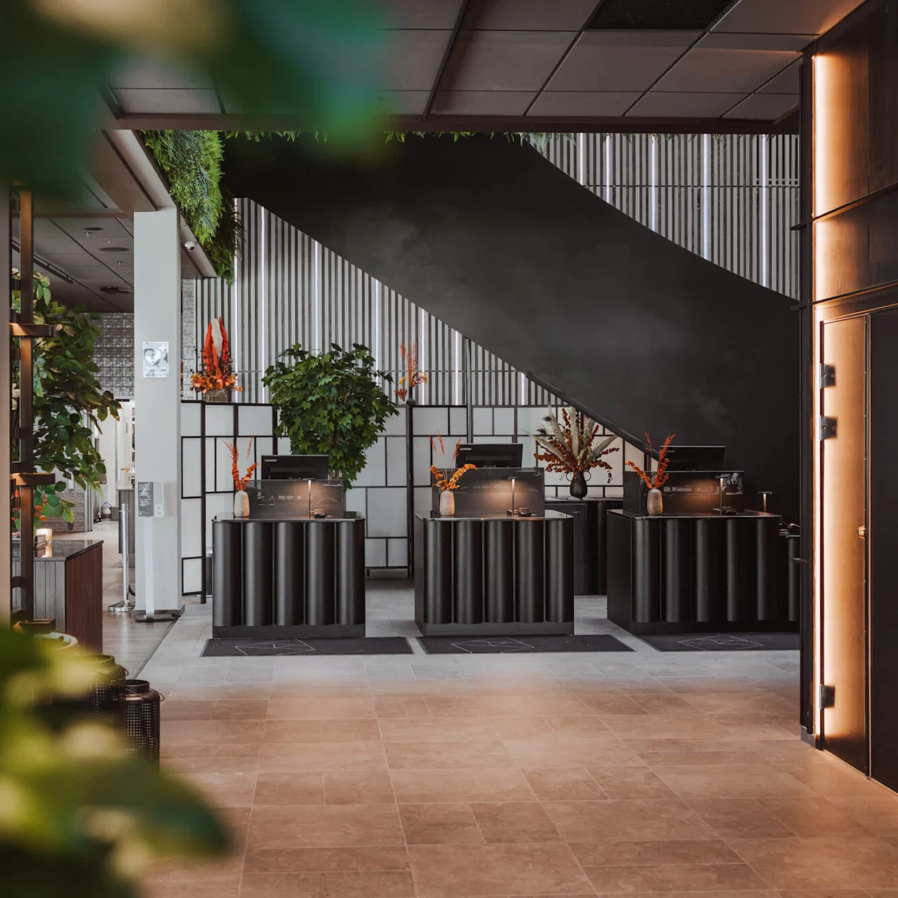 Receptiondisken med datorer står under en modern trappa, omgiven av växter och blomsterarrangemang, vilket skapar en inbjudande atmosfär i en samtida lobby.