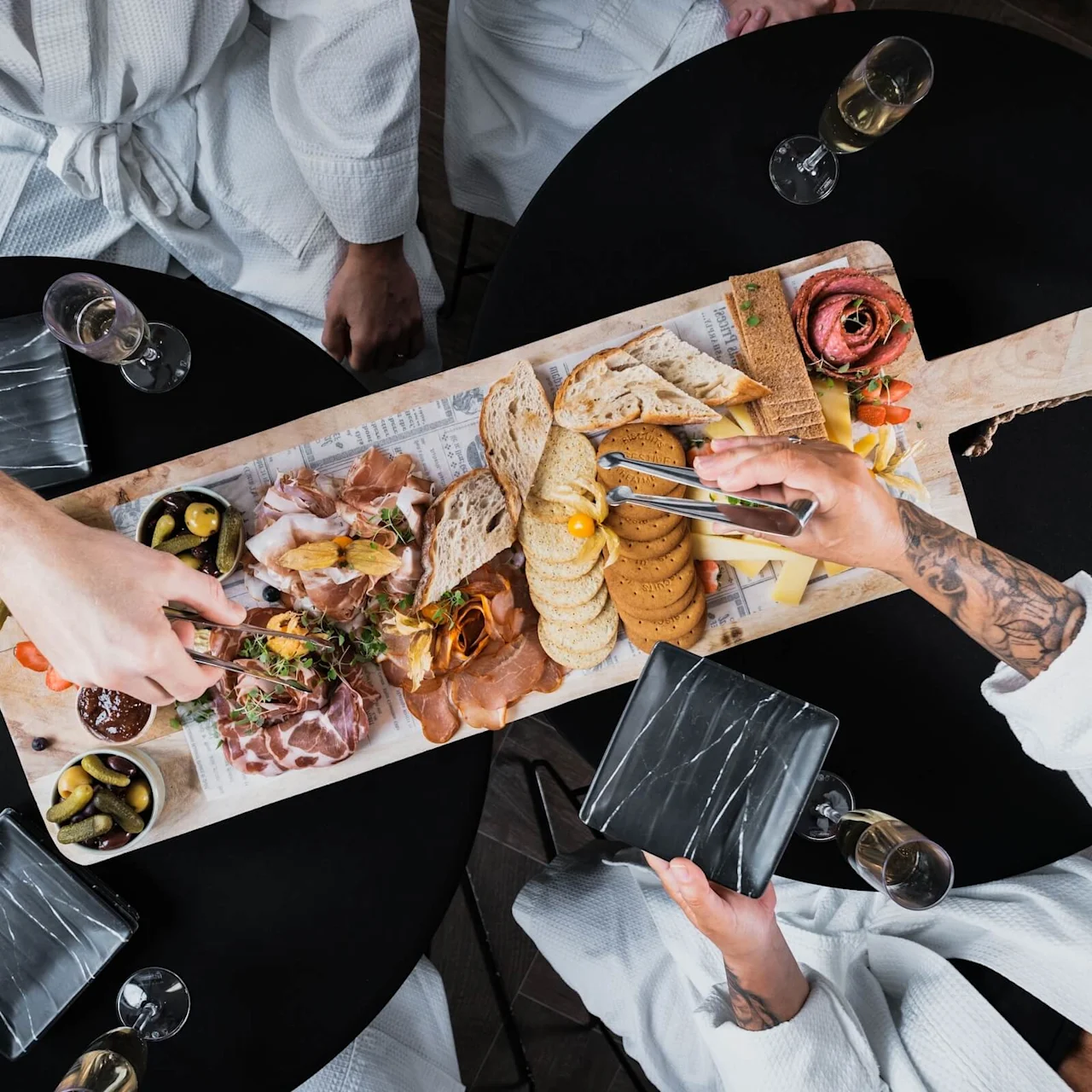 A wooden serving board holds assorted cheeses, cured meats, bread, and olives. Two people in white robes use utensils to serve themselves at a black table with glasses of champagne.
