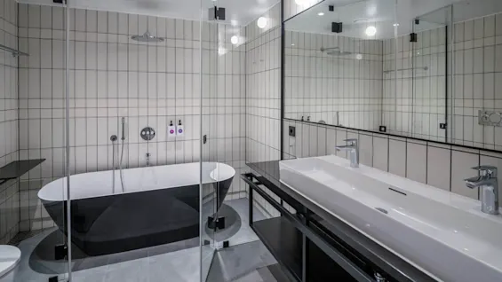 Et moderne bad har et stort sort badekar, omgitt av glasspaneler, med hvite og grå flisevegger. En lang hvit vask med to kraner står overfor.