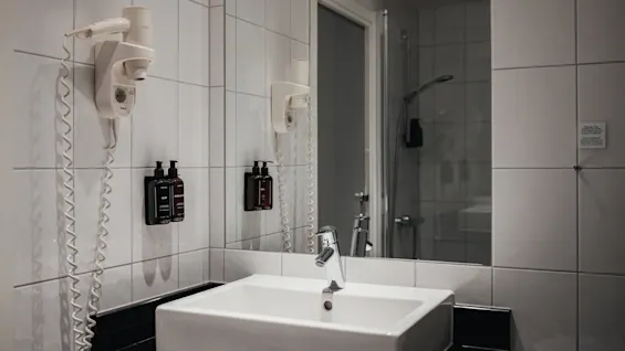Et moderne badeværelse med hvid vask, stort spejl, vægmonteret hårtørrer og sæbedispensere. En bruser er synlig i spejlbilledet.