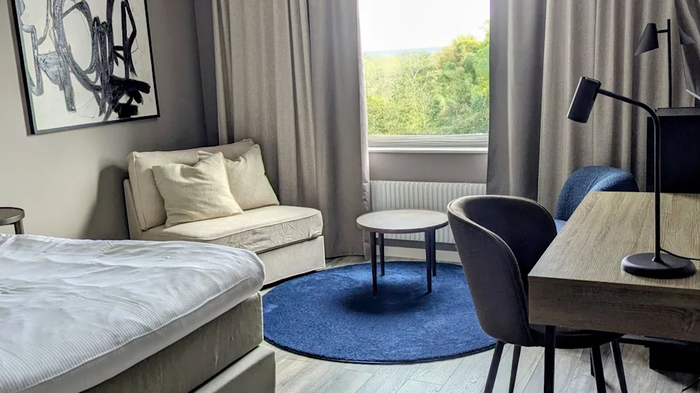 Ett modernt hotellrum med en bekväm säng, en mysig sittgrupp och ett skrivbord, med en fridfull utsikt över grönskande natur utanför fönstret.