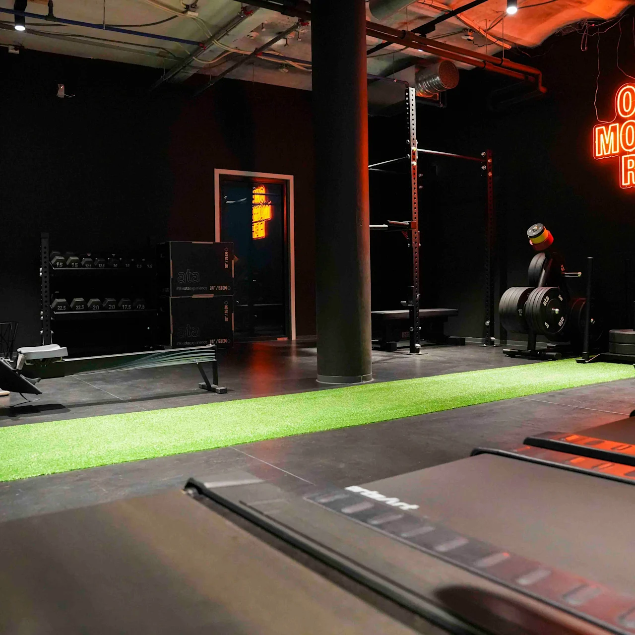 Gyminteriør med treningsutstyr som tredemølle, romaskin og vekter. Neonskilt på veggen med teksten "ONE MORE REP." Mørke vegger og tak med industrielle elementer.