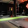 Gyminteriør med treningsutstyr som tredemølle, romaskin og vekter. Neonskilt på veggen med teksten "ONE MORE REP." Mørke vegger og tak med industrielle elementer.