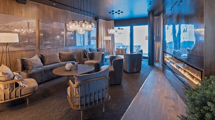 A modern living room features plush gray furniture around a wooden table, ambient spherical lights hang from the ceiling, with a cozy fireplace along a wood-paneled wall.