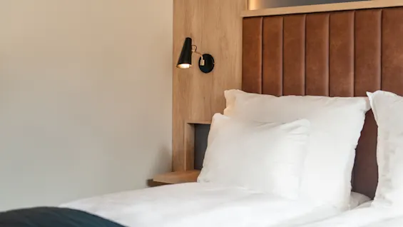 En hyggelig hotelseng med hvide puder, en brun polstret sengegavl og en stilfuld sort væglampe.