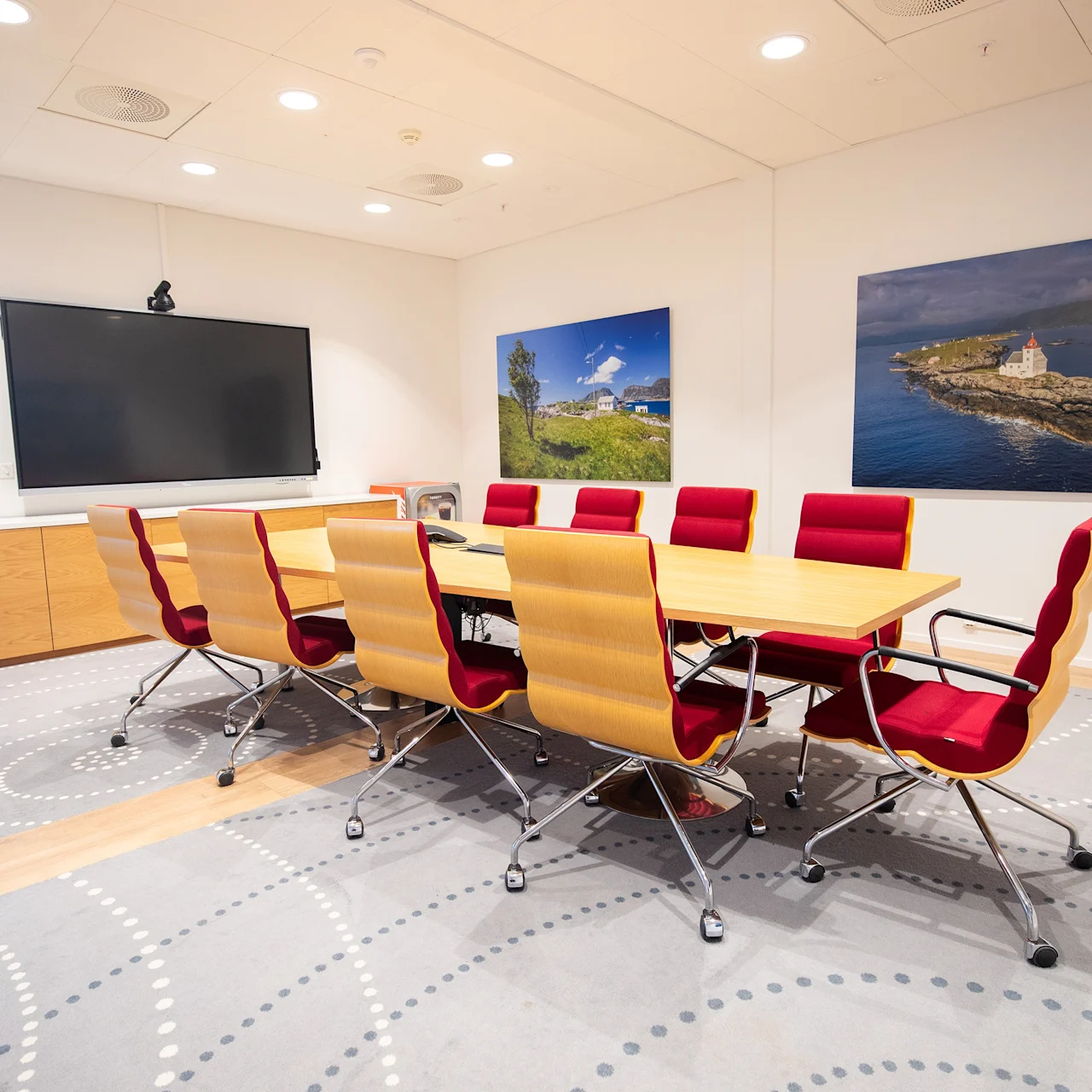 Et konferanserom med et rektangulært bord omgitt av stilige stoler. En stor skjerm er montert på veggen, og et skap står under. Landskapsbilder pryder veggene.