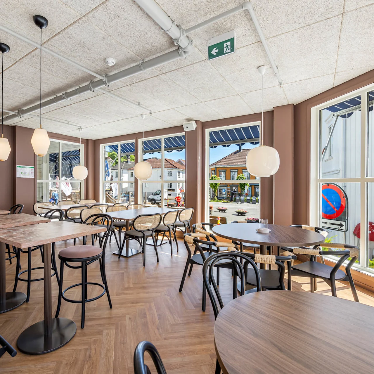 A modern cafe interior with empty tables and chairs, pendant lights, and large windows overlooking a street.