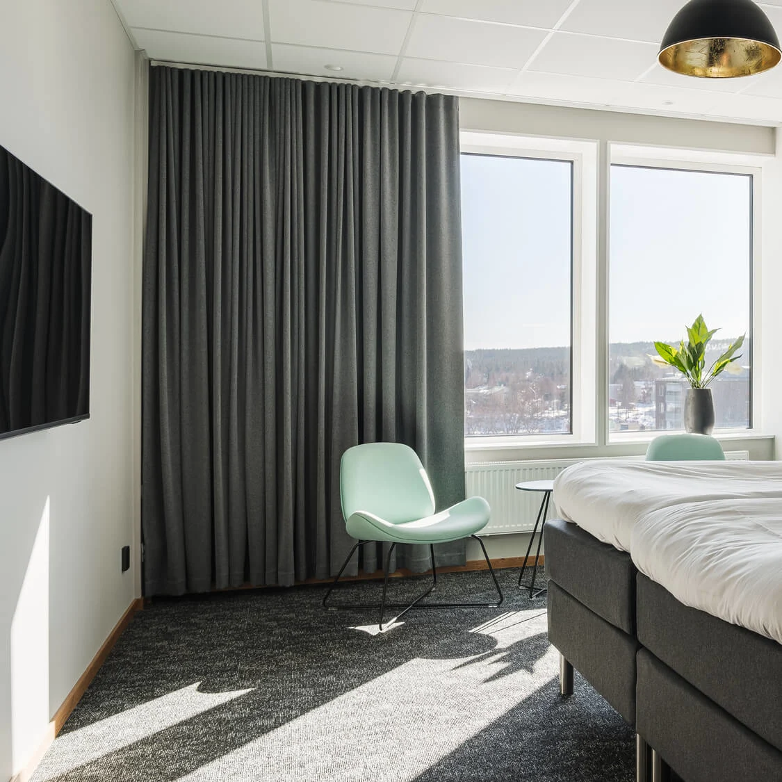 En prydligt bäddad säng står i ett ljust rum. Solljus strömmar in genom stora fönster med utsikt över ett snöigt landskap. Nära finns en grön stol som kompletterar den moderna inredningen.