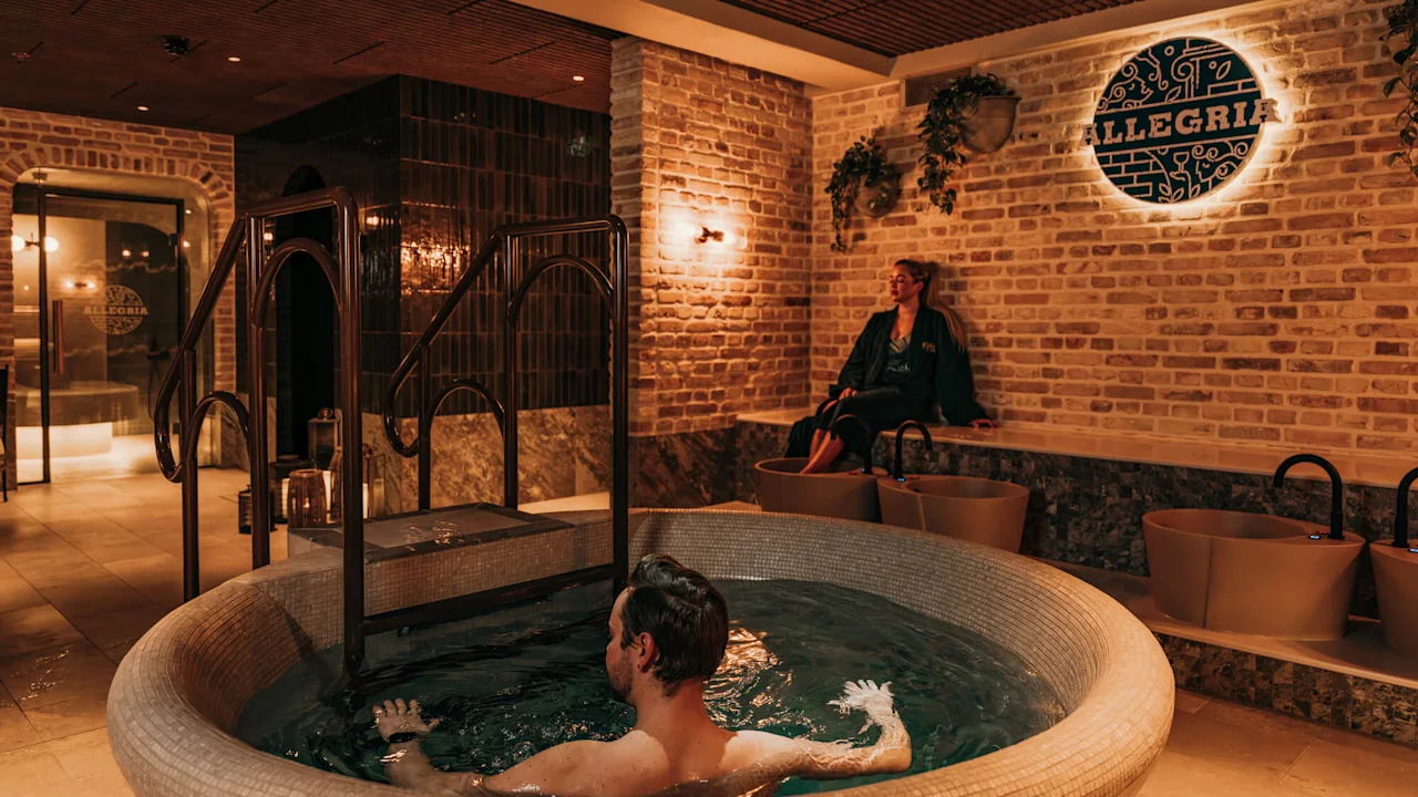 A man relaxes in a circular indoor spa pool with warm lighting, while a woman sits nearby. A sign reads "ALLEGRIA" on a brick wall.