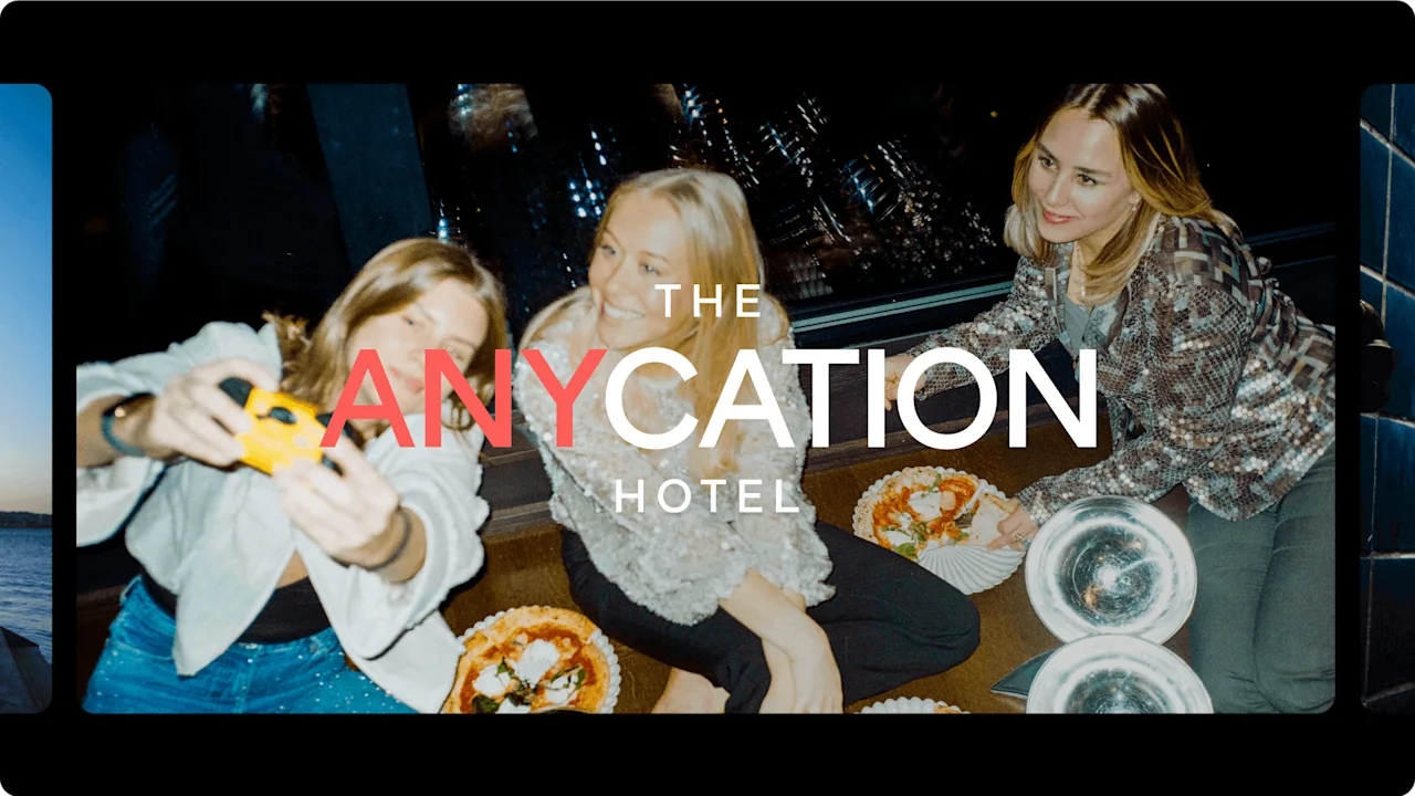 Kolme ihmistä istuu sisällä, ottaa selfietä ja nauttii pizzasta. Tekstissä lukee "THE ANYCATION HOTEL".