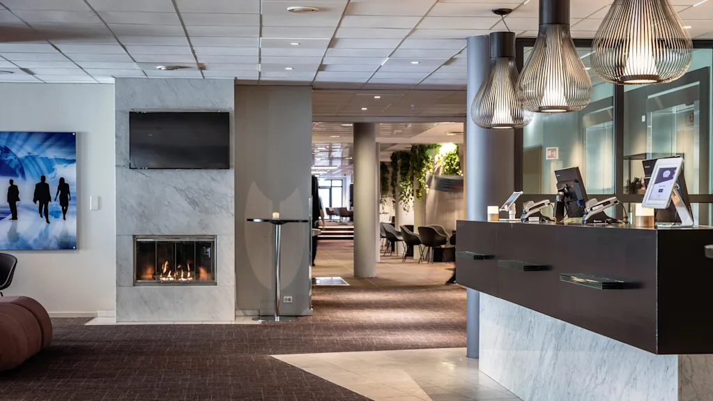 Reception desk situated in a modern hotel lobby with computers on it; nearby, a lit fireplace under a TV, and a hallway leading to seating areas accented by greenery.