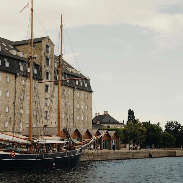 En historisk kanalscene med stor mursteinsbygning, seilbåt fortøyd og annen båt i det fjerne. Folk langs vannkanten.