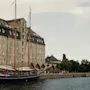 En historisk kanalscene med stor mursteinsbygning, seilbåt fortøyd og annen båt i det fjerne. Folk langs vannkanten.