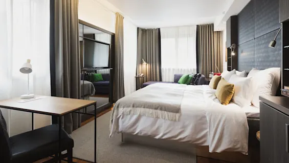 En pænt redt seng med hvide lagner og gule puder i et moderne hotelværelse. Et lille skrivebord med lampe står i nærheden, og gardiner indrammer store vinduer.