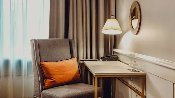 A gray armchair with an orange cushion beside a desk with a phone and lamp, against a curtained window in a warmly lit room.