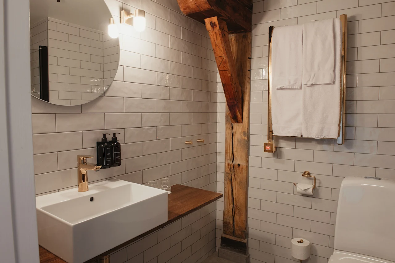 Et moderne badeværelse med hvide fliser, en vask med guldfarvede armaturer og en rustik træbjælke. Hvide håndklæder er pænt foldet.