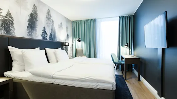 A neatly made bed with white linens stands against a wallpapered wall with tree motifs. A wooden desk and chair sit by a window with teal curtains; a TV is mounted opposite.