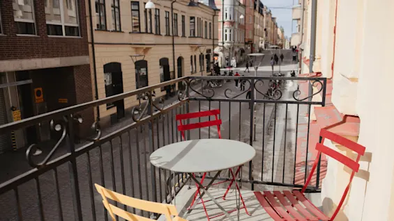 Ett runt bord och tre färgglada stolar står på en balkong med utsikt över en kullerstensgata med historiska byggnader och fotgängare.