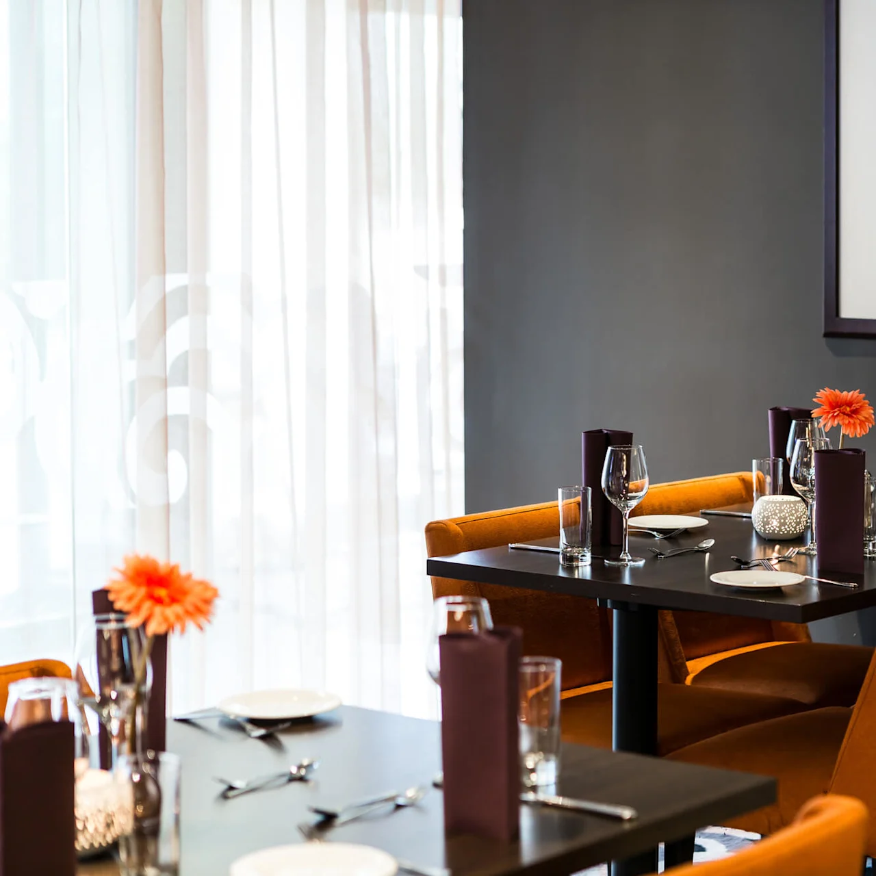 Table set for dining with glasses, plates, and napkins; adorned with orange flowers in a dimly lit restaurant; portrait on wall.