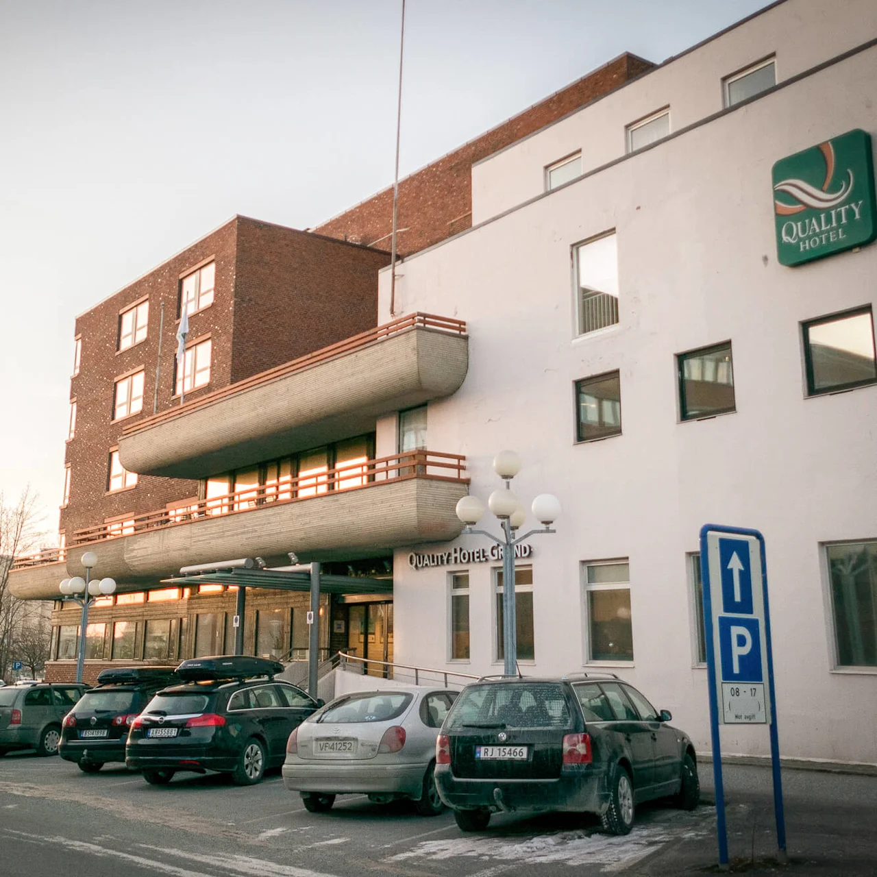 A hotel with a "Quality Hotel" sign on the facade, cars parked in front, and a blue parking sign. The sky is softly lit, suggesting sunrise or sunset.