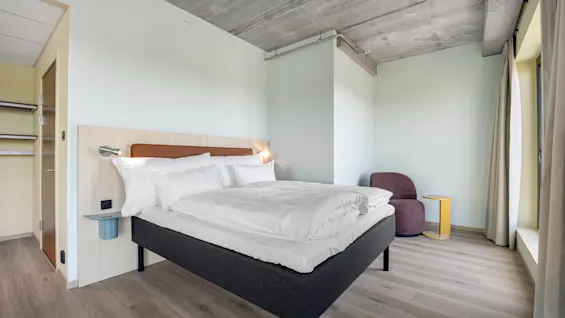 A modern and inviting hotel room with a comfortable bed, a cozy chair, and a window letting in natural light.