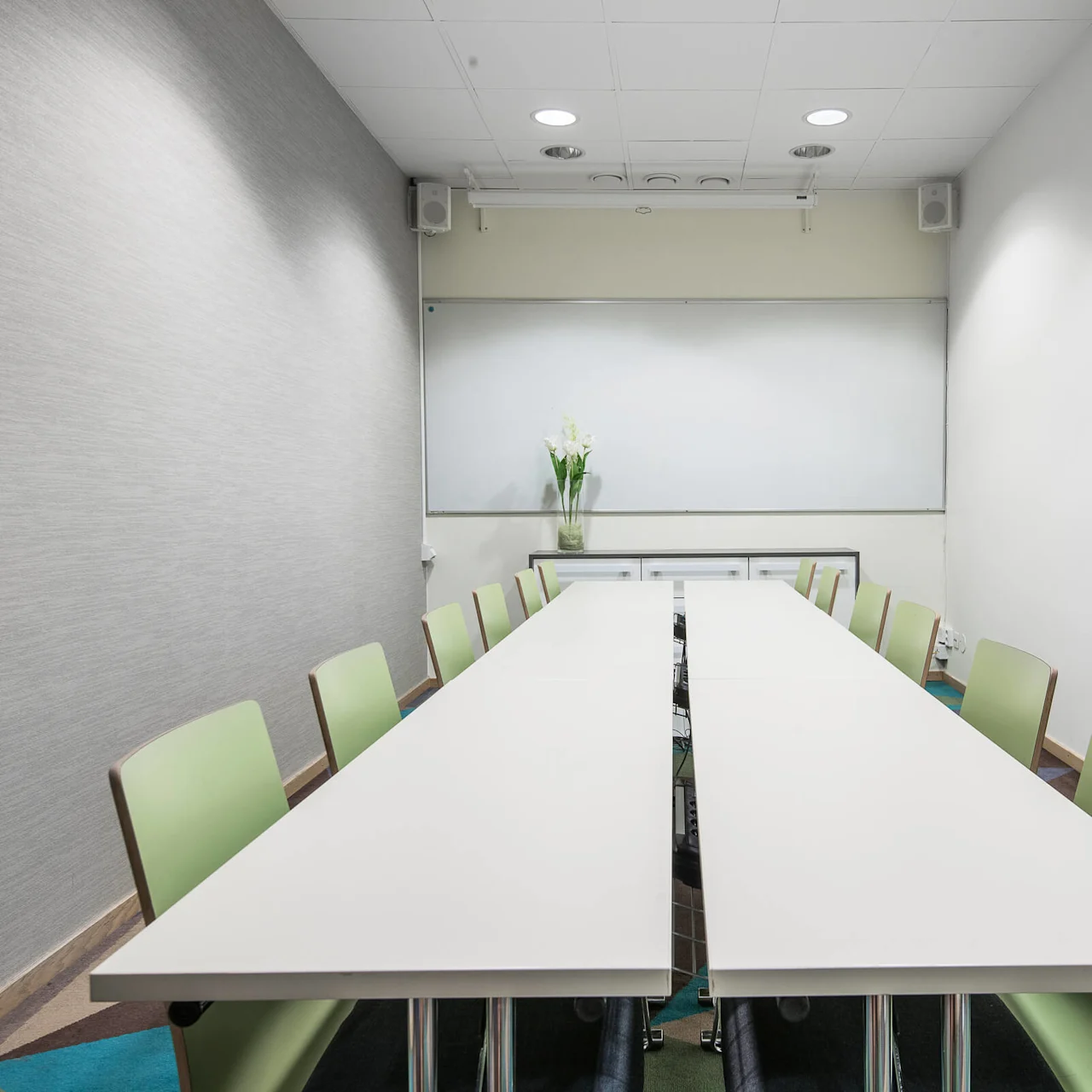 Konferensbord omgivet av gröna stolar, beläget i ett konferensrum med en whiteboard på väggen och en blomvas.