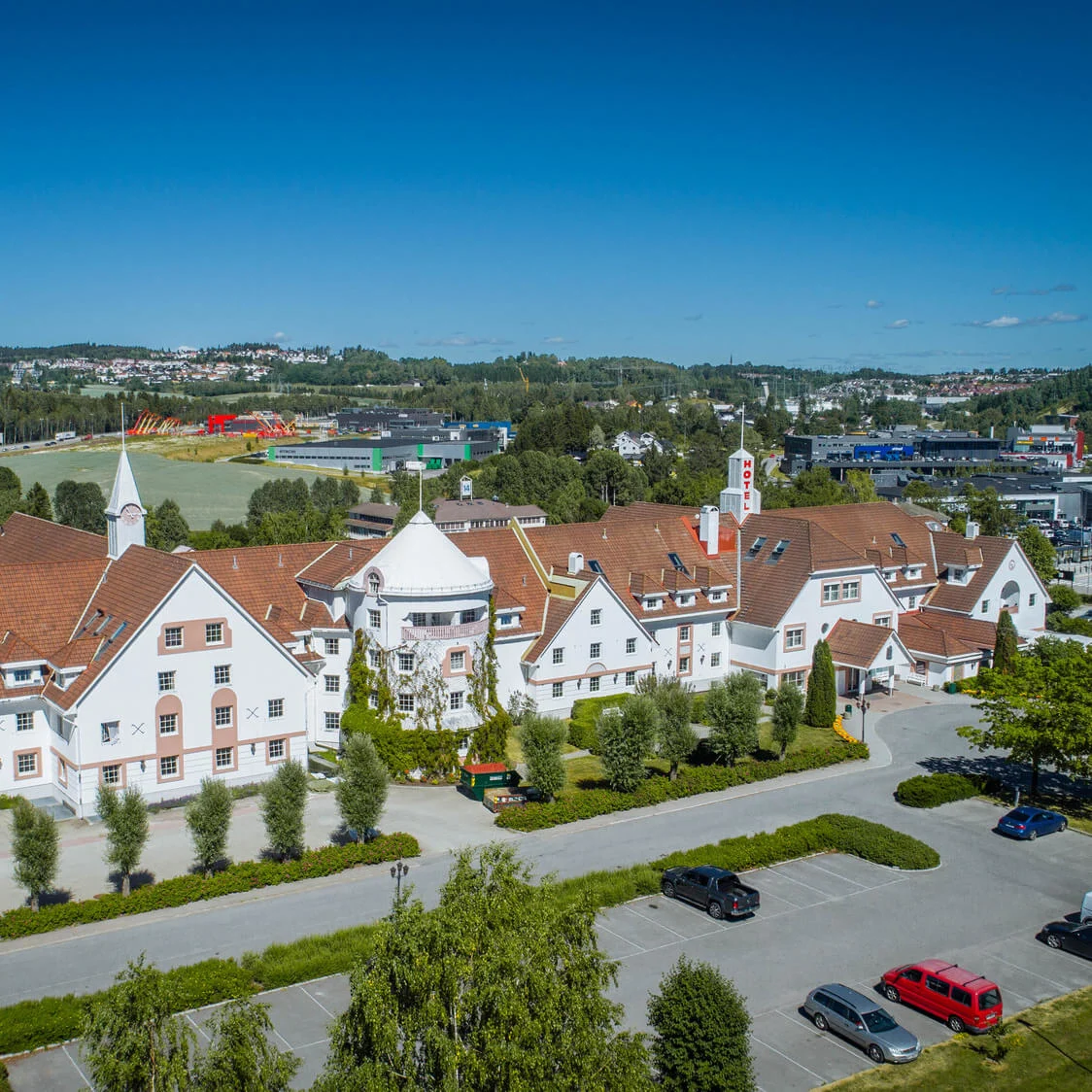 Hotel med røde tegltage står, omgivet af parkering og grønt, beliggende i et frodigt landskab under en klar blå himmel. Synlig tekst: "Quality Airport Hotel Værnes”.