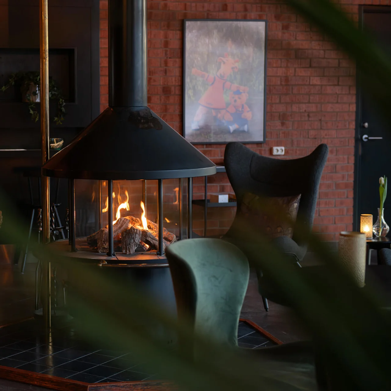 Fireplace radiates warmth, surrounded by chairs in a cozy room with exposed brick walls. A basket of firewood and a framed picture provide rustic ambiance.