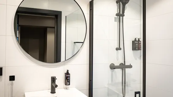 A modern bathroom with white tiled walls, a round mirror above a sink, and a shower area with dark fixtures.