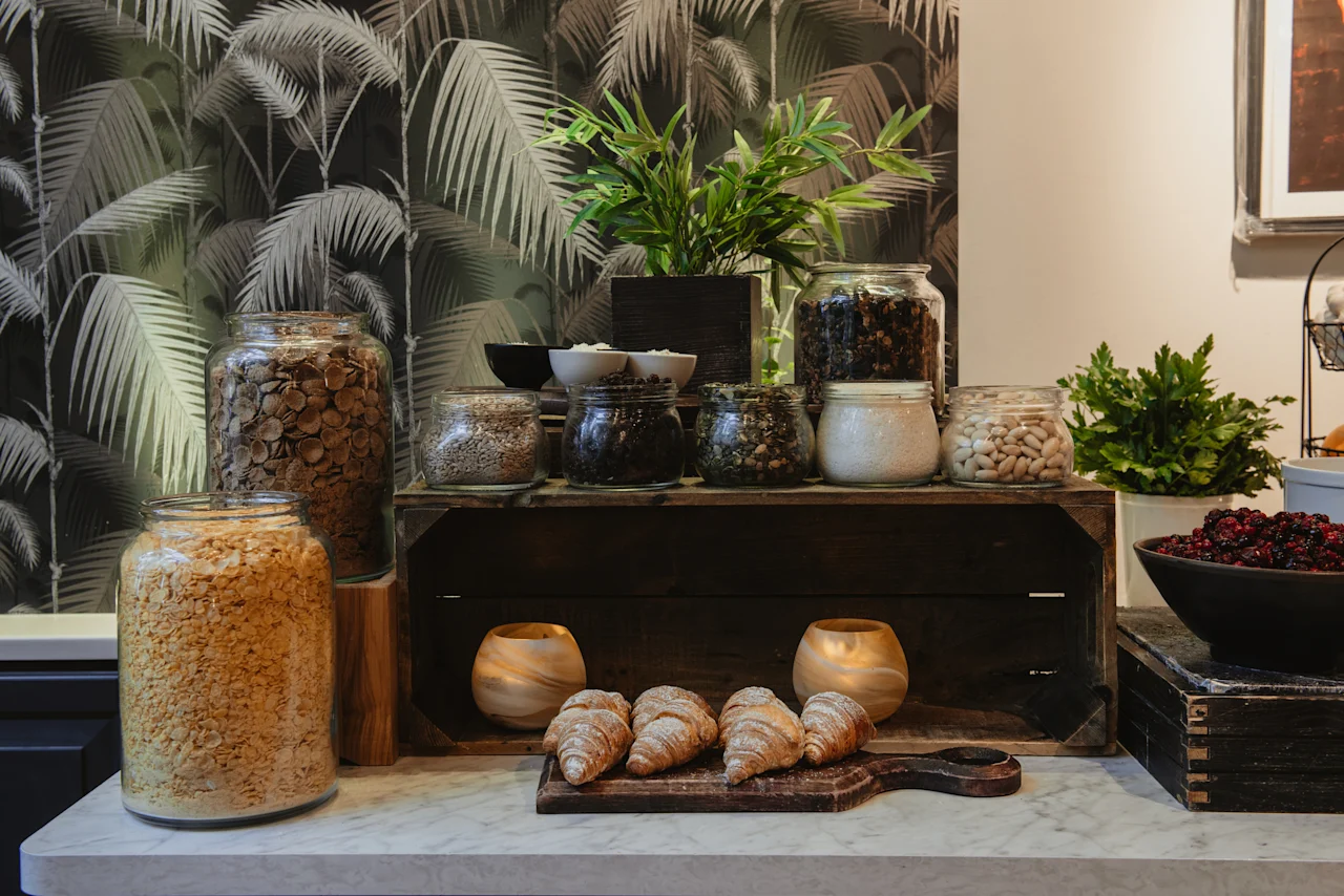 En morgenmadsbuffet med krukker med morgenmadsprodukter, nødder og tørret frugt, samt friske croissanter på et træbræt.