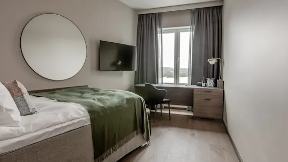 Et pænt indrettet hotelværelse med en seng dækket af et grønt tæppe. Et rundt spejl hænger over, et TV er vægmonteret, og et vindue har udsigt over et landskab.
